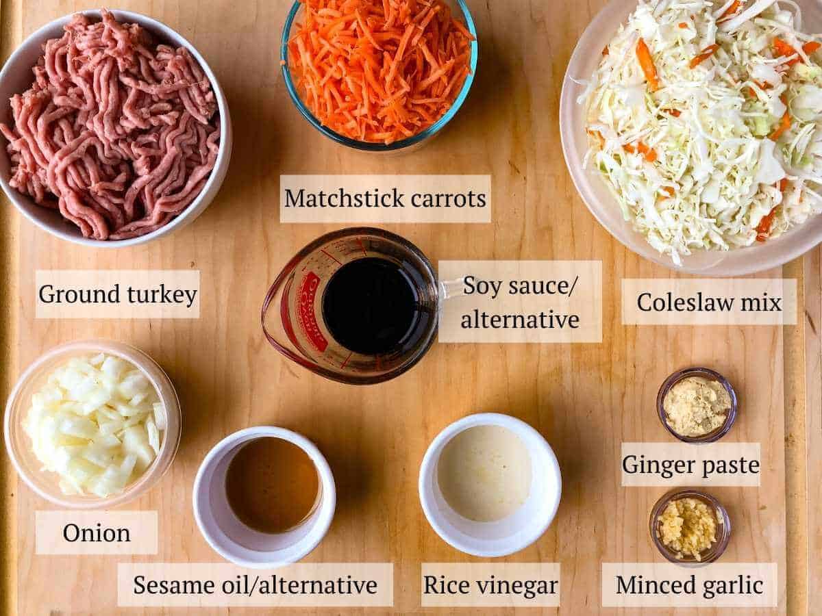 The ingredients for inside out egg rolls including ground turkey, matchstick carrots, coleslaw mix, soy sauce or alternative, ginger paste, minced garlic, sesame oil or alternative, and chopped onion.