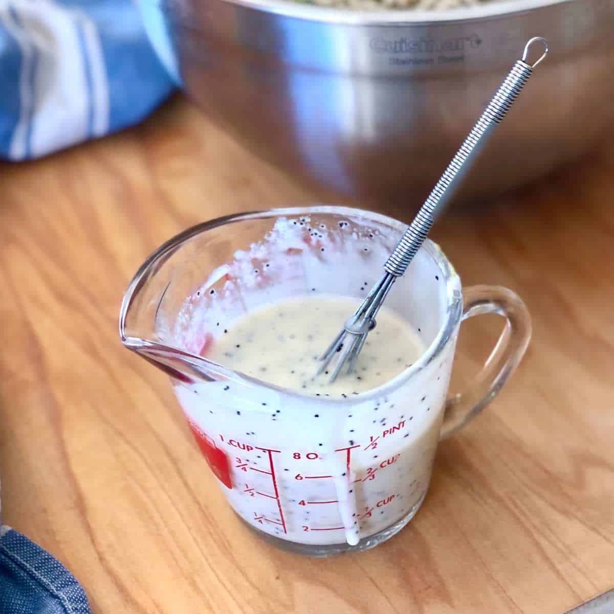 A glass measuring cup with vegan poppy seed dressing and a whisk.