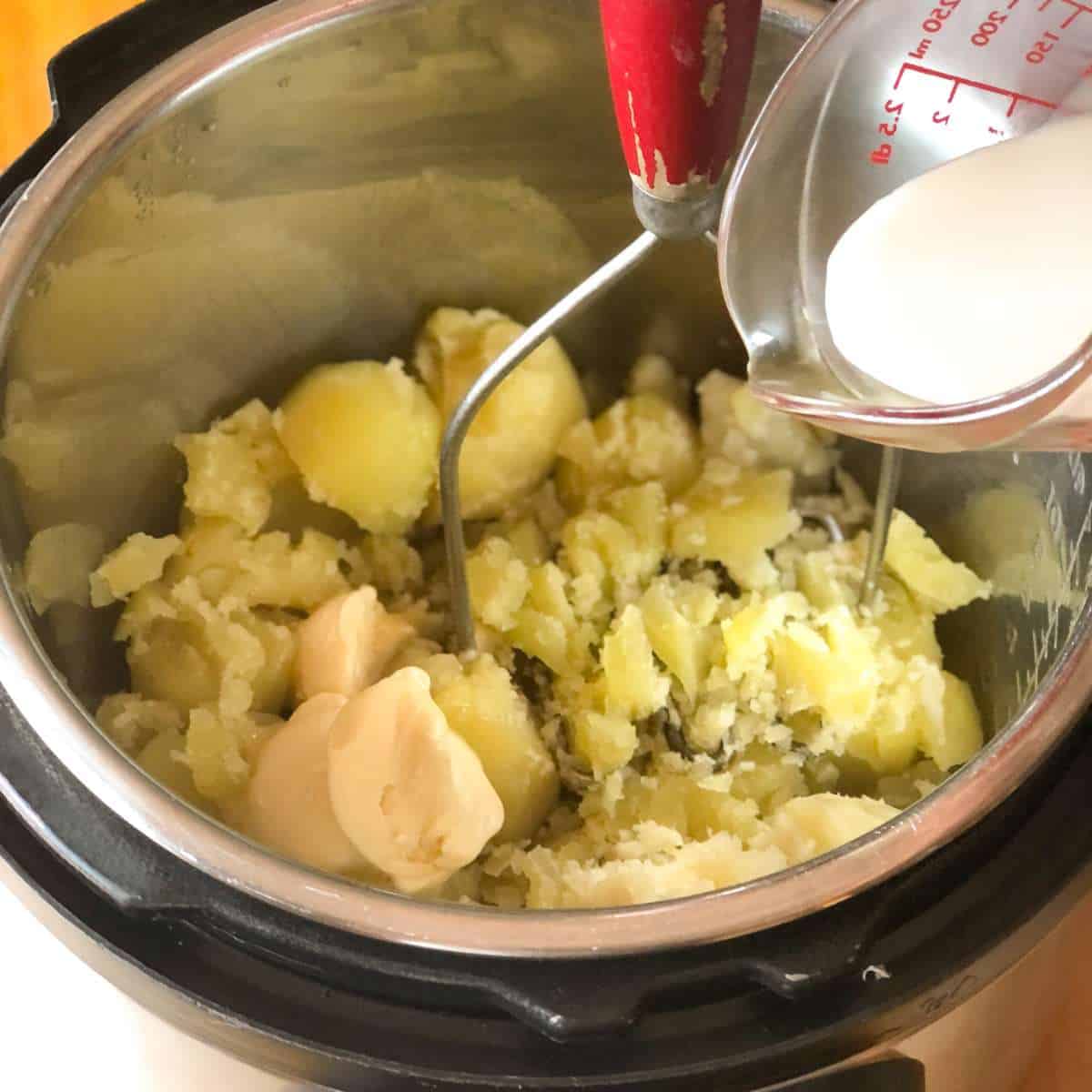 Mashed potatoes with dairy free margarine in a pot with a glass measuring cup of dairy free milk being poured onto the potatoes.